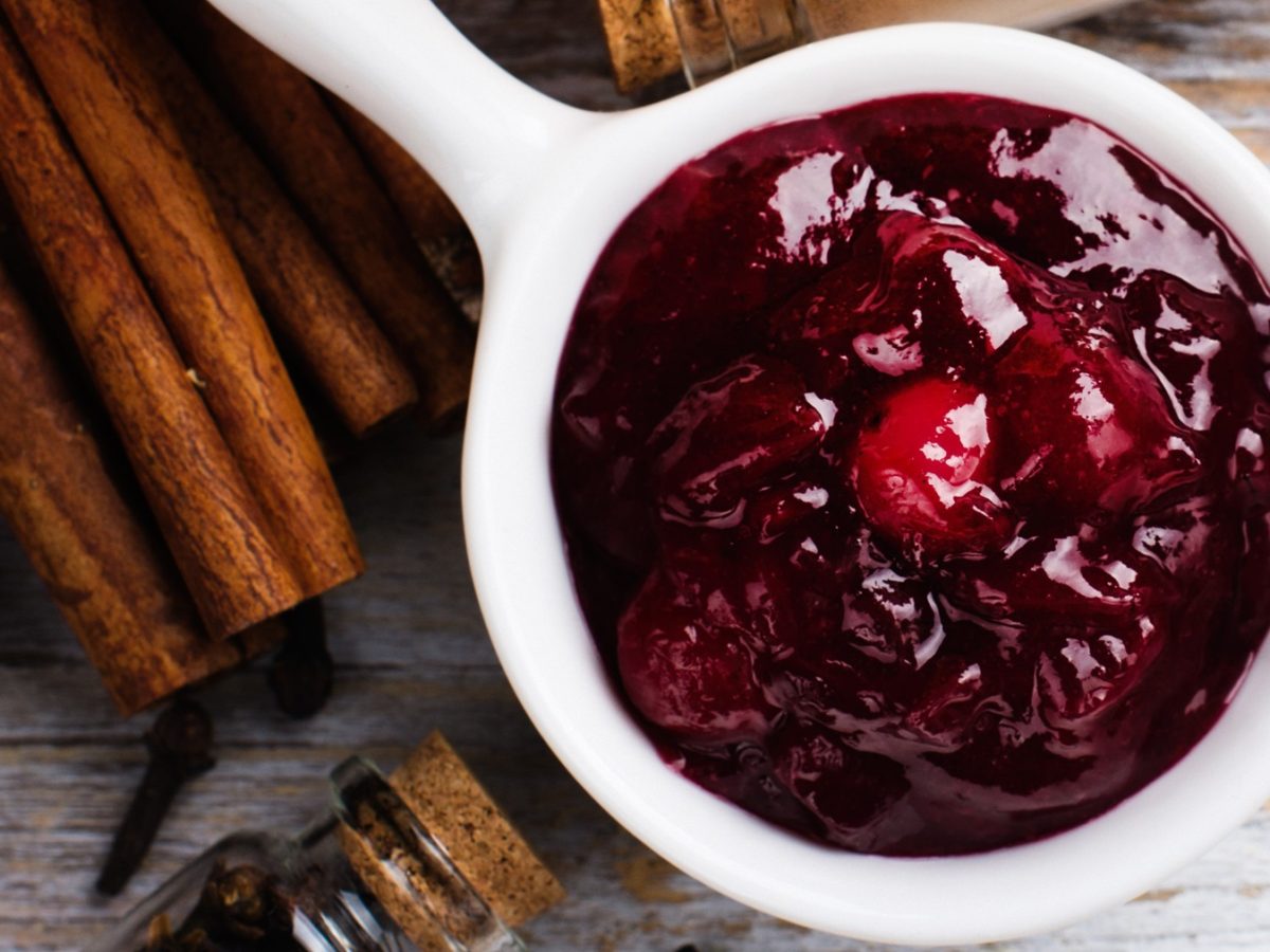 Eine kleine weiße Sauciere mit den Pfefferkirschen auf einem Holztisch mit allerlei Gewürzen von oben fotografiert.