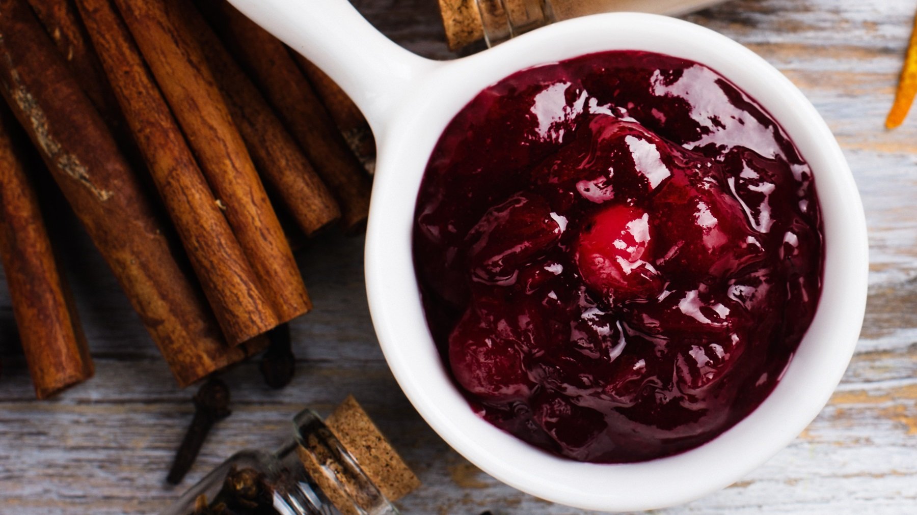 Eine kleine weiße Sauciere mit den Pfefferkirschen auf einem Holztisch mit allerlei Gewürzen von oben fotografiert.