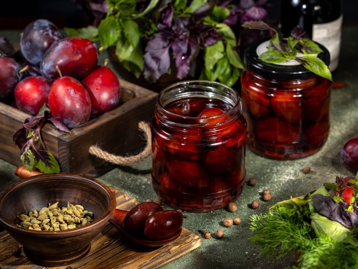 Nahaufnahme von eingekochten Pflaumen in Glas, umgeben von Kardamom, Piment, frischen Kräutern und prallen, dunklen Früchten.