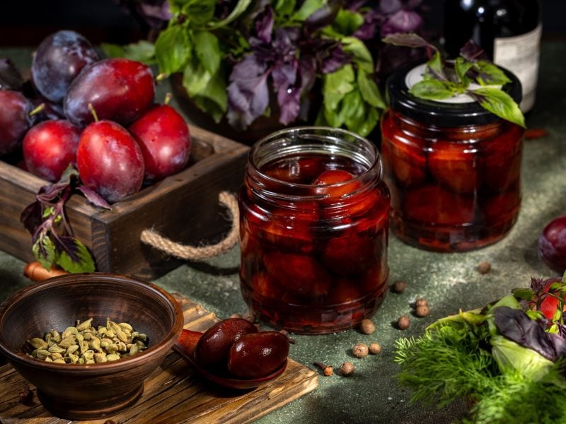 Nahaufnahme von eingekochten Pflaumen in Glas, umgeben von Kardamom, Piment, frischen Kräutern und prallen, dunklen Früchten.