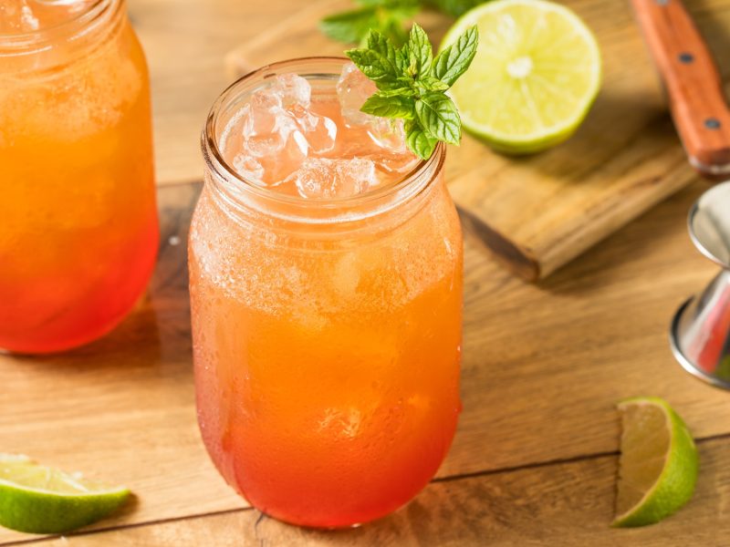 Zwei Gläser Planters Punch mit Eiswürfeln auf Holzplatte, vorn garniert mit Minze, dahinter Limetten und Barzubehör.