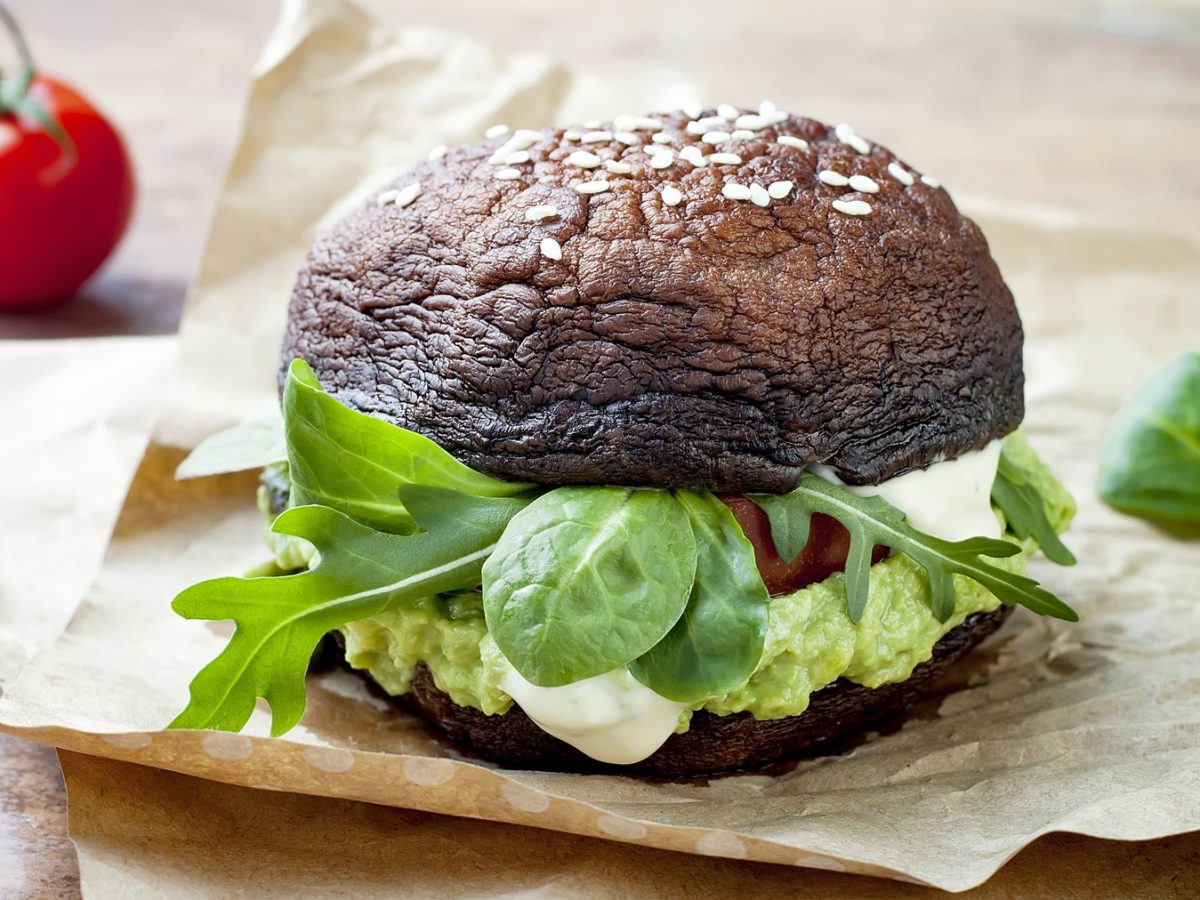Der Portobello-Burger auf einem dunklen Holztisch mit Tomaten und Basilikum im Hintergrund.