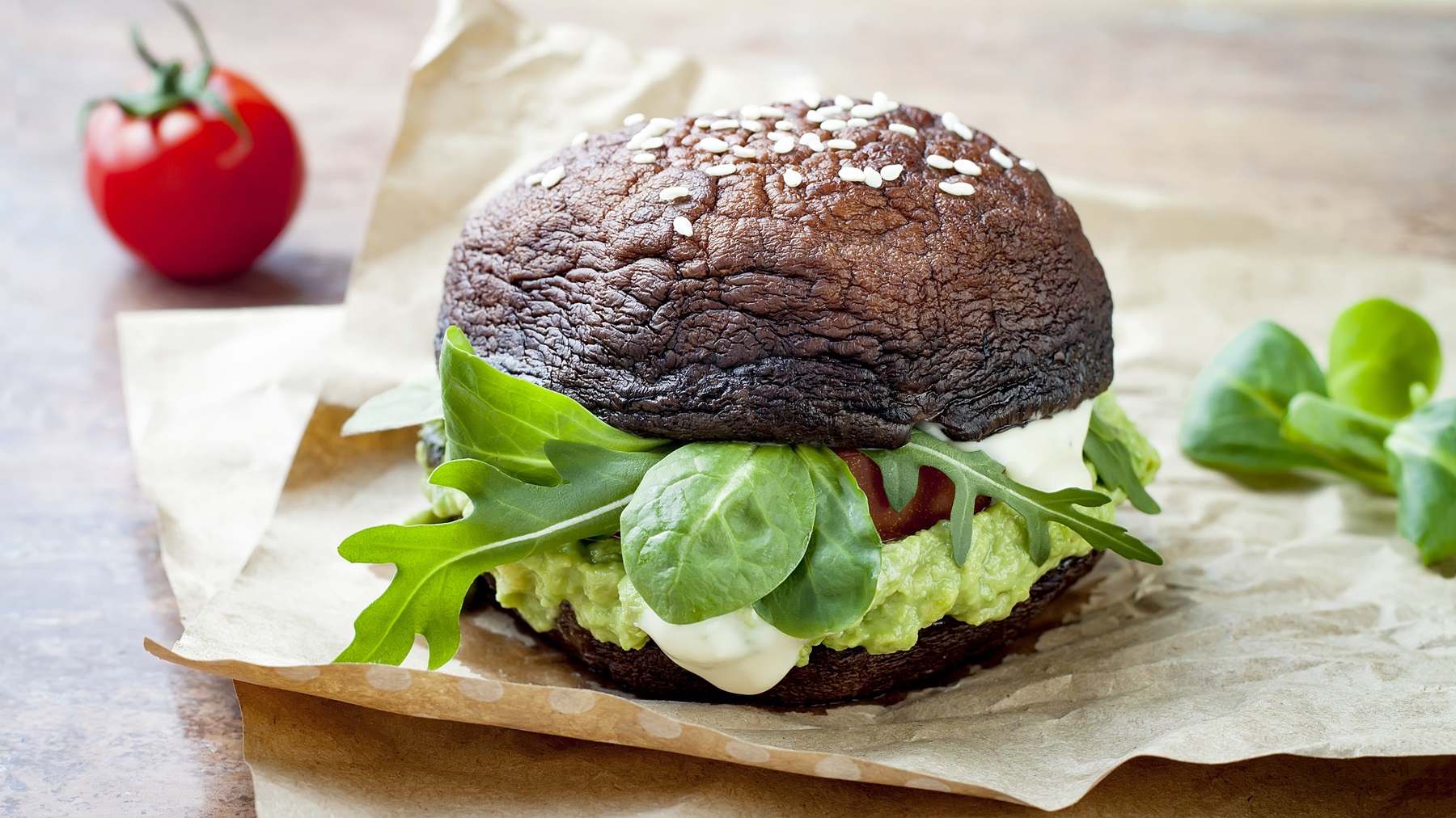 Der Portobello-Burger auf einem dunklen Holztisch mit Tomaten und Basilikum im Hintergrund.
