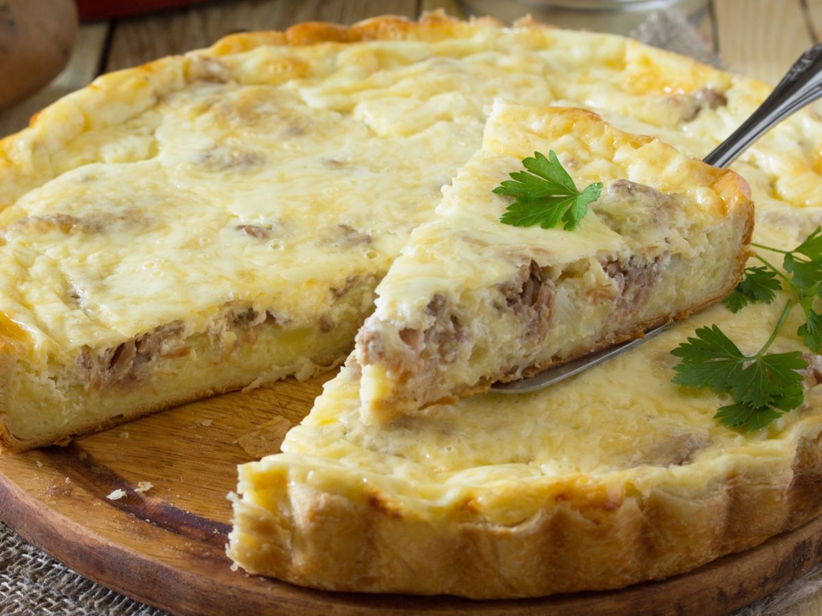 Die Quiche auf einem hellen Tisch von der Seite fotografiert. Ein Stück wurde mit dem Tortenheber bereits hochgehoben.