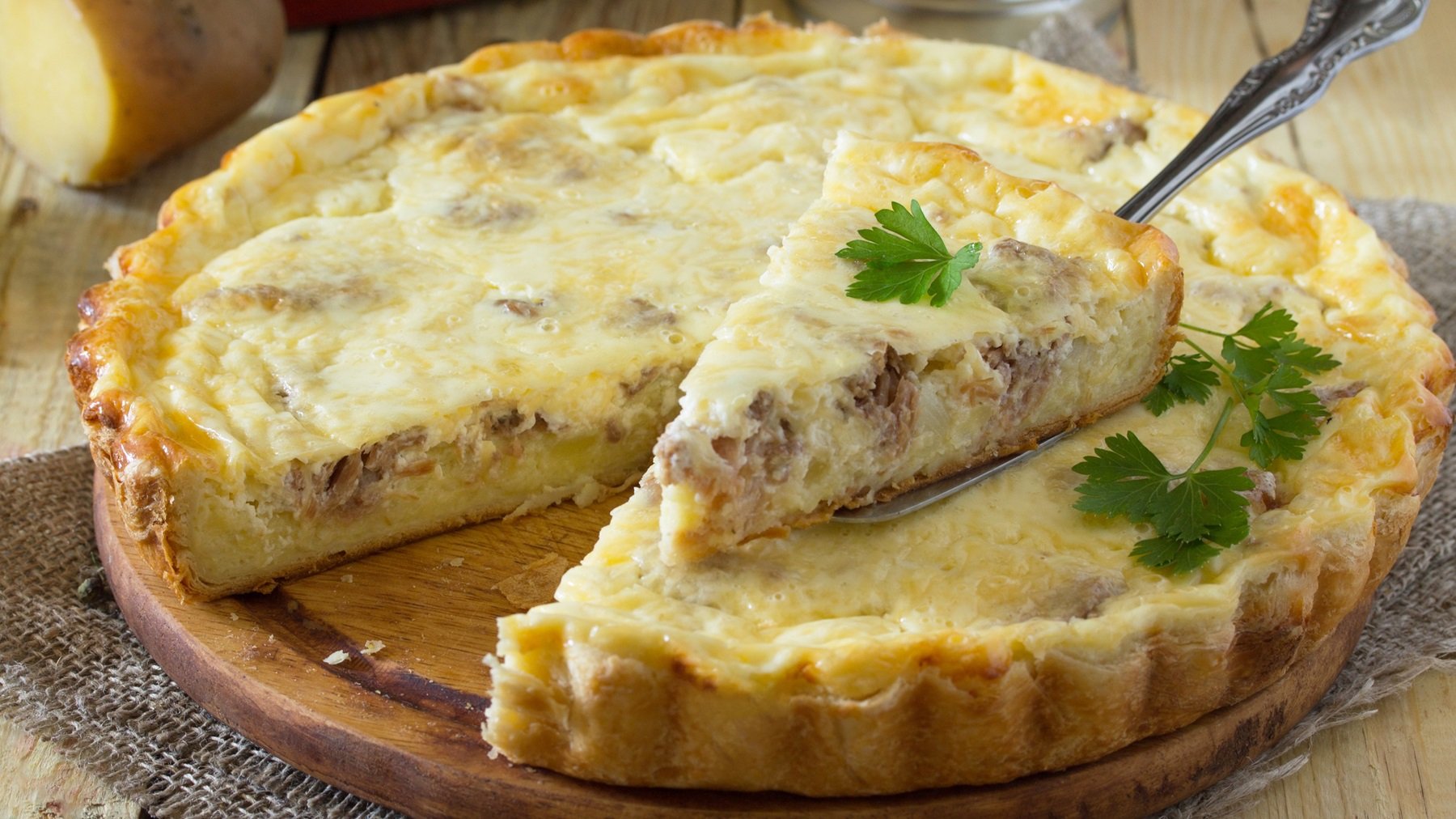 Die Quiche auf einem hellen Tisch von der Seite fotografiert. Ein Stück wurde mit dem Tortenheber bereits hochgehoben.