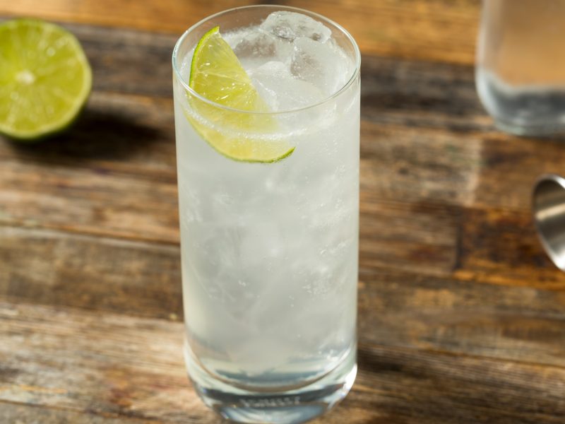 Erfrischender Ranch Water Cocktail mit Limettenscheibe und Eis in hohem Glas auf einem verwitterten Holztisch mit Barzubehör.