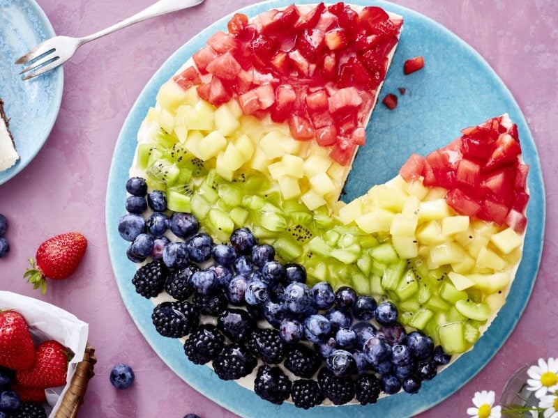 Eine runde Torte mit buntem Obstbelag in Regenbogenfarben liegt auf einem blauen Teller. Die Torte ist in Streifen mit Erdbeeren, Melone, Kiwi, Heidelbeeren und Brombeeren belegt. Ein Stück wurde herausgeschnitten und liegt auf einem kleinen Teller daneben. Um die Torte herum liegen frische Beeren und ein kleiner Korb mit Erdbeeren und Heidelbeeren auf einer rosa Tischdecke. Im Vordergrund sind weiße Gänseblümchen zu sehen.