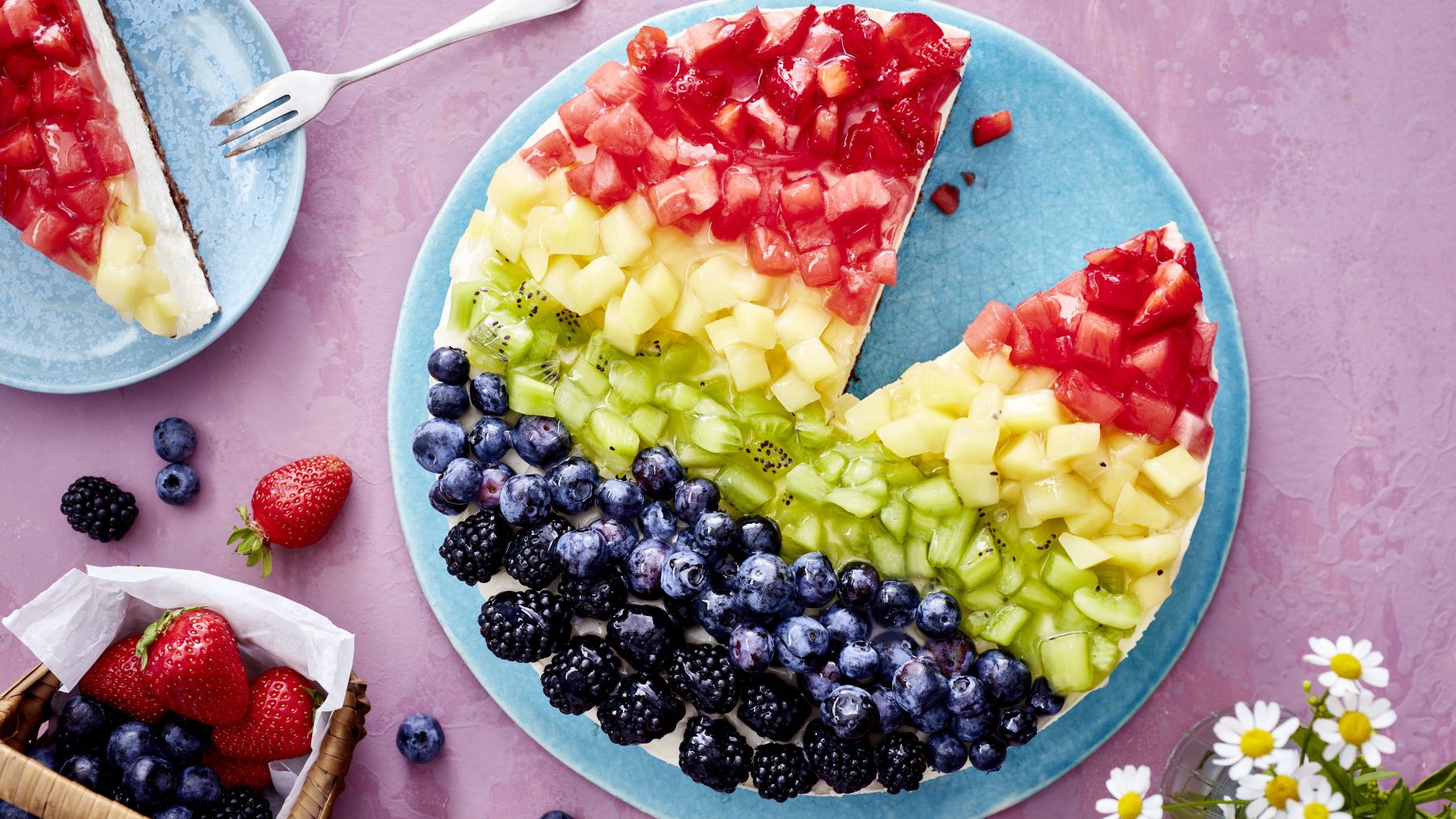 Eine runde Torte mit buntem Obstbelag in Regenbogenfarben liegt auf einem blauen Teller. Die Torte ist in Streifen mit Erdbeeren, Melone, Kiwi, Heidelbeeren und Brombeeren belegt. Ein Stück wurde herausgeschnitten und liegt auf einem kleinen Teller daneben. Um die Torte herum liegen frische Beeren und ein kleiner Korb mit Erdbeeren und Heidelbeeren auf einer rosa Tischdecke. Im Vordergrund sind weiße Gänseblümchen zu sehen.