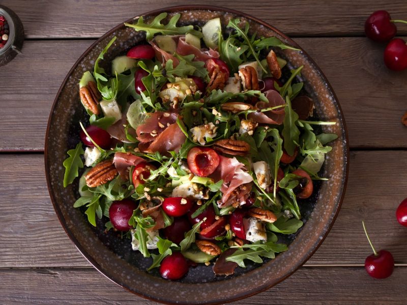 Sommerlicher Salatteller mit Rucola, Kirschen, Blauschimmelkäse, Nüssen und Schinken auf dunkler Keramikschale.