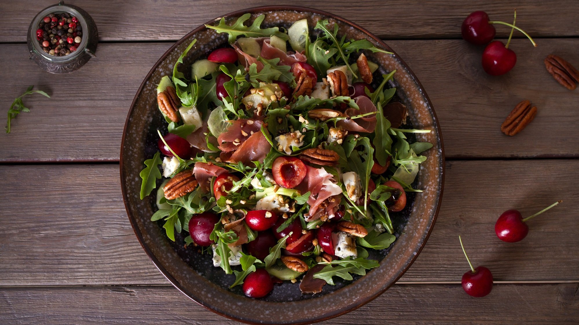 Sommerlicher Salatteller mit Rucola, Kirschen, Blauschimmelkäse, Nüssen und Schinken auf dunkler Keramikschale.