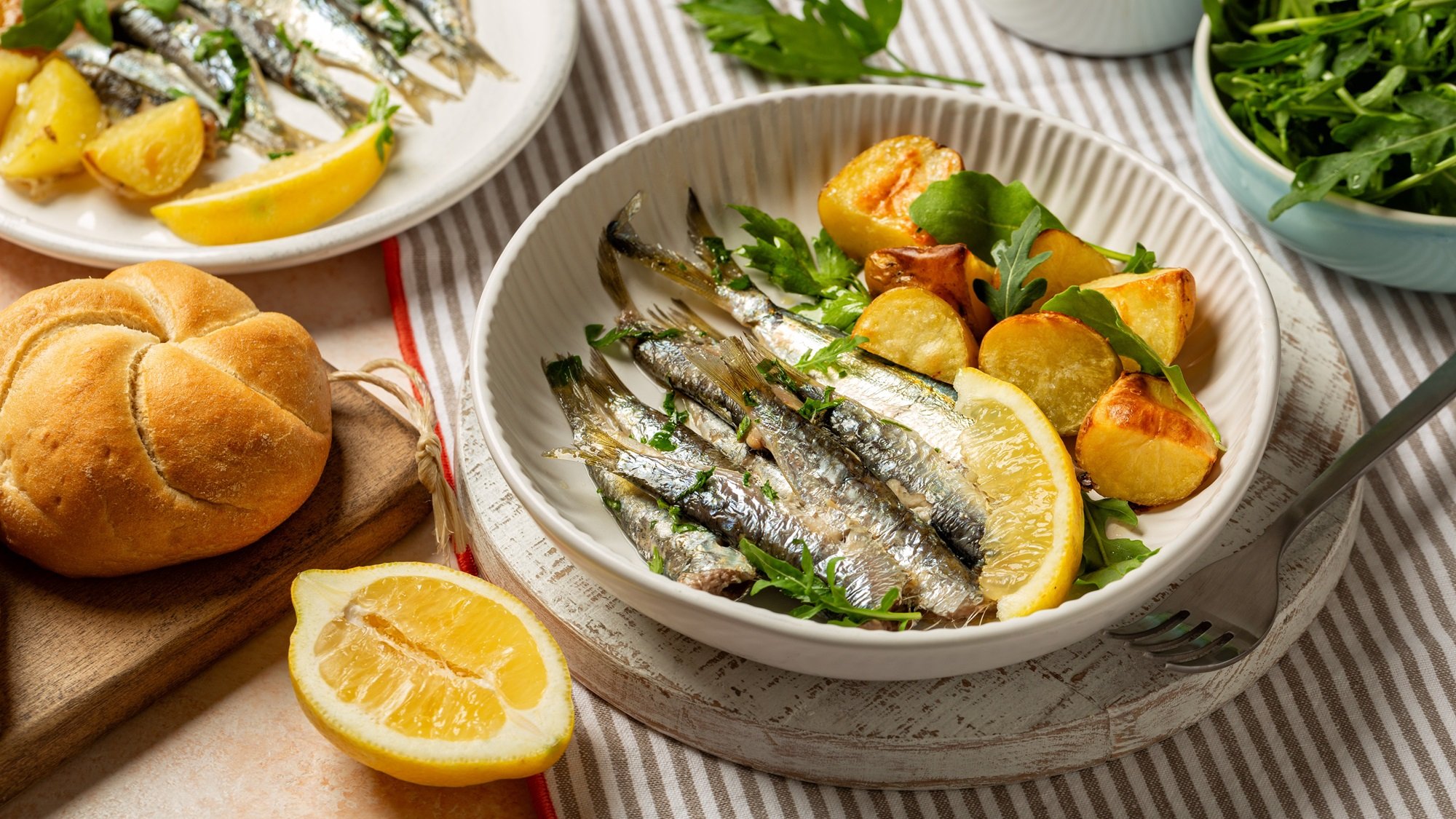 Appetitlich angerichtete Sardinen mit Kräutern und Zitronenstücken auf Rucola, serviert mit Kartoffeln auf gestreifter Tischdecke.