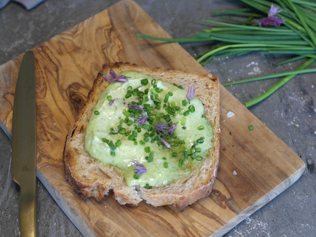 Eine Scheibe Brot mit der grünen Schnittlauchbutter auf einem Holzbrett und steinernem Untergrund. Alles von schräg oben fotografiert.