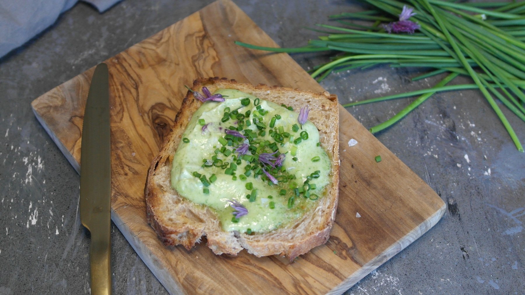 Eine Scheibe Brot mit der grünen Schnittlauchbutter auf einem Holzbrett und steinernem Untergrund. Alles von schräg oben fotografiert.