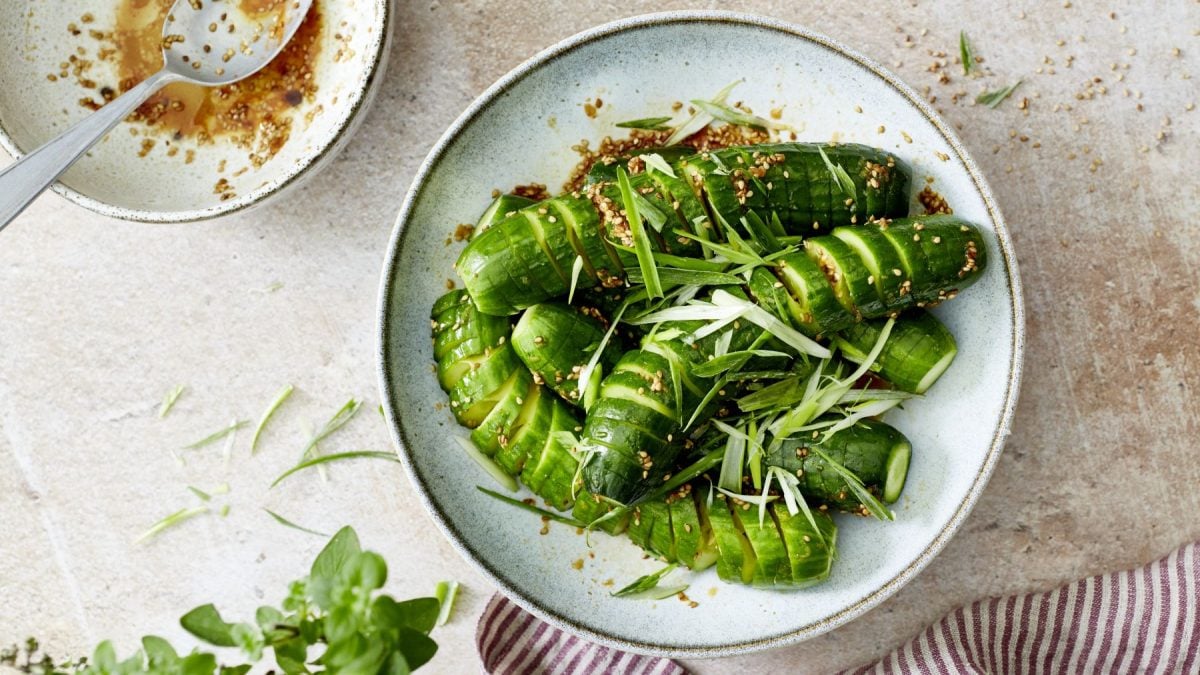 In einer hellen, schlichten Keramikschale liegen Verschärfte Spirelligurken wie beim Korean BBQ, die mit goldbraunem Sesam und feinen Streifen von Frühlingszwiebeln bestreut sind. Die Gurken glänzen in einer würzigen Marinade, daneben steht eine Schale mit etwas restlicher Sauce und einem Löffel. Im Vordergrund sind Kräuter und ein gestreiftes Tuch zu sehen, der Hintergrund ist hell und minimalistisch gehalten.
