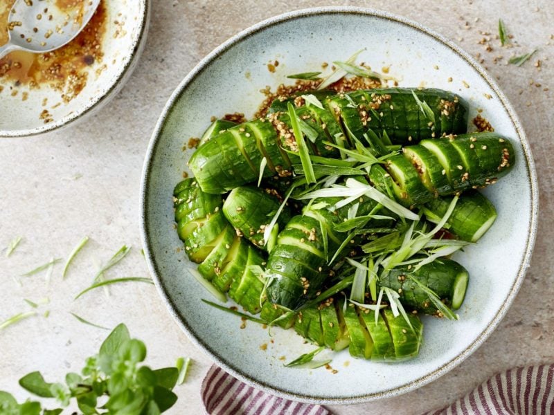 In einer hellen, schlichten Keramikschale liegen Verschärfte Spirelligurken wie beim Korean BBQ, die mit goldbraunem Sesam und feinen Streifen von Frühlingszwiebeln bestreut sind. Die Gurken glänzen in einer würzigen Marinade, daneben steht eine Schale mit etwas restlicher Sauce und einem Löffel. Im Vordergrund sind Kräuter und ein gestreiftes Tuch zu sehen, der Hintergrund ist hell und minimalistisch gehalten.