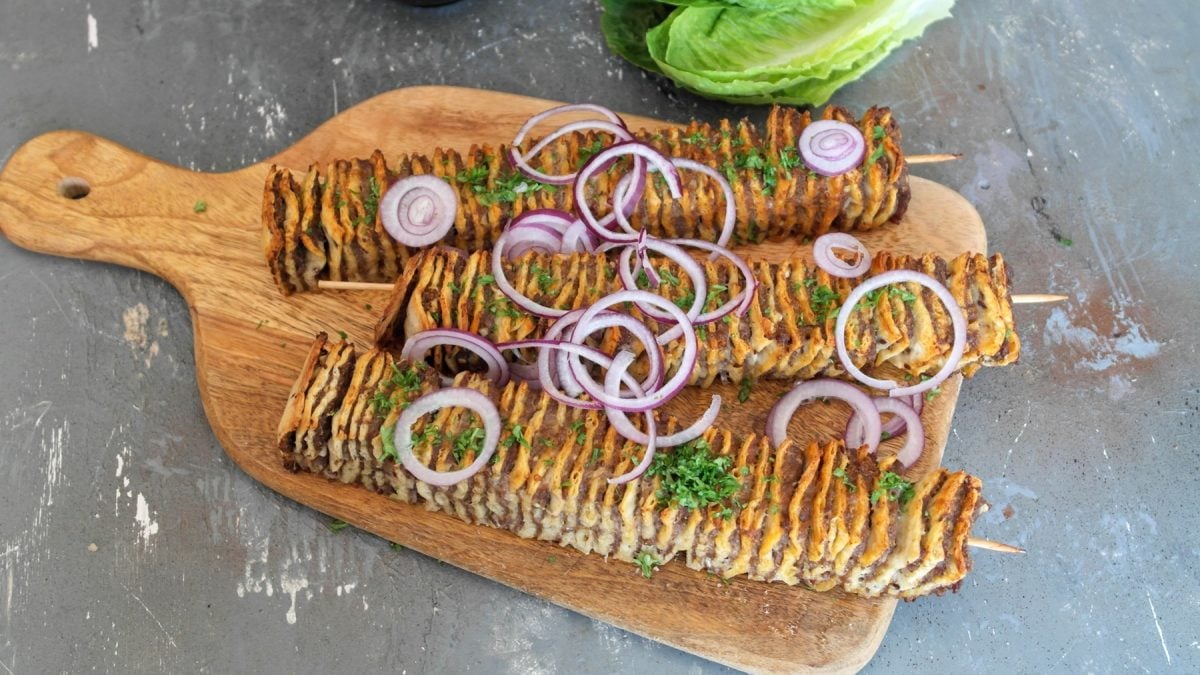 Drei Kebap-Spieße auf einem Holzbrett, mit Zwiebeln und Petersilie garniert. Alles direkt von oben fotografiert.