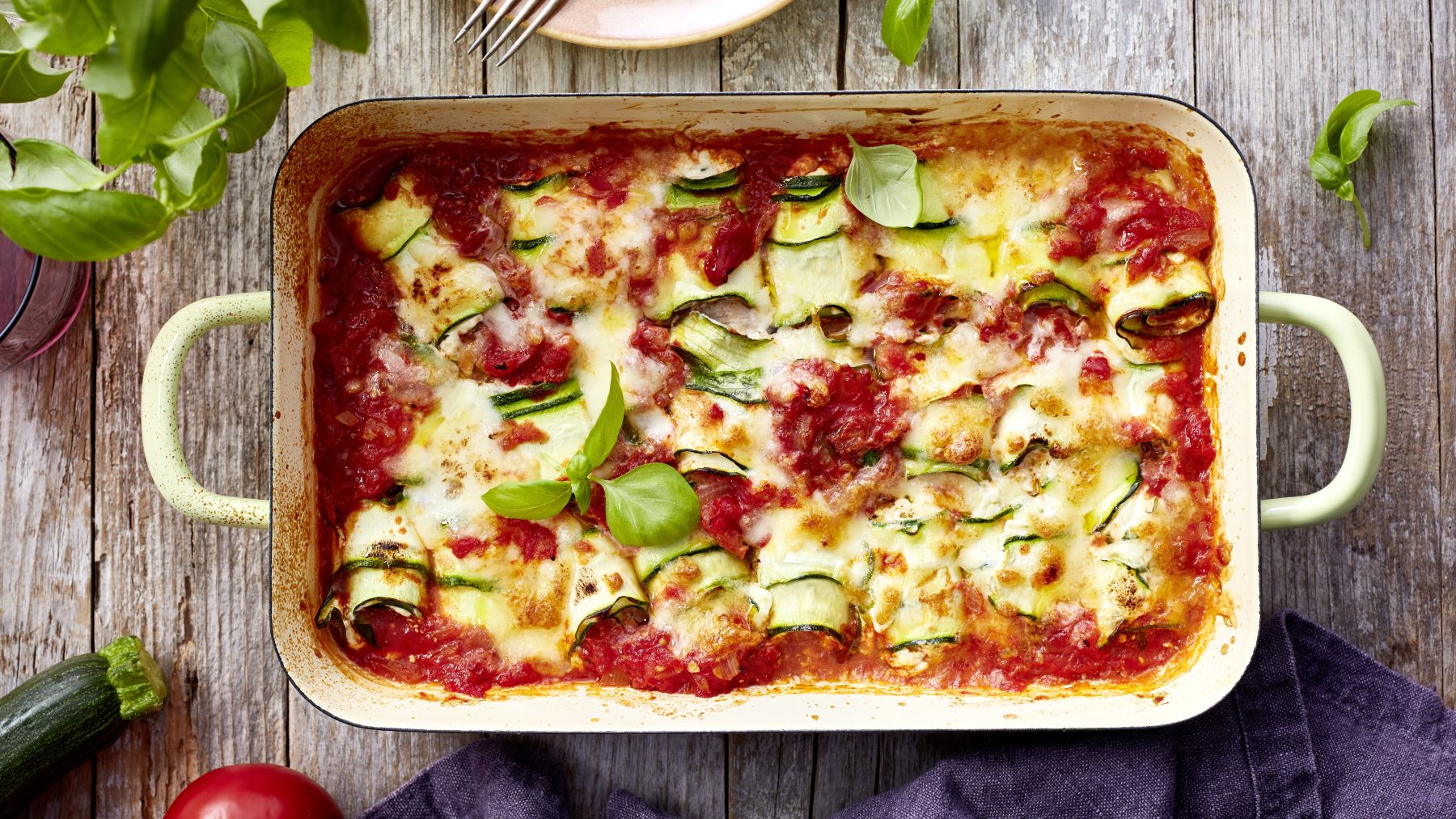 Ein rechteckiger Auflauf steht auf einem rustikalen Holztisch. Der Auflauf besteht aus geschichteten Zucchinistreifen, Ricotta und einer tomatigen Sauce, überbacken mit geschmolzenem Käse. Einige frische Basilikumblätter garnieren das Gericht. Am Rand sind eine Zucchini, eine Tomate und ein Glas zu sehen. Das Bild vermittelt einen frischen, mediterranen Eindruck und lädt zum Genießen ein.