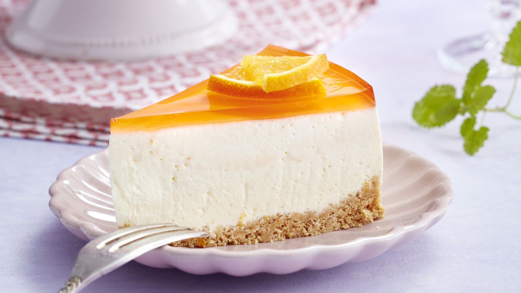 Ein Stück Aperol-Spritz-Torte mit Keksboden, weißer Frischkäsecreme und orangefarbenem Guss, garniert mit Orangenscheiben auf rosafarbenem Kuchenteller.