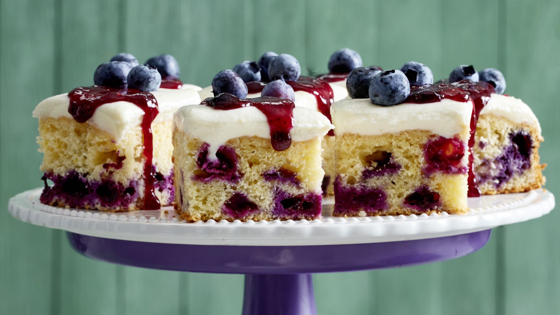 Saftige Stücke Heidelbeer-Zitronenkuchen mit cremigem Topping, fruchtiger Beerensoße und frischen Heidelbeeren, angerichtet auf einer Tortenplatte vor grünem Hintergrund.