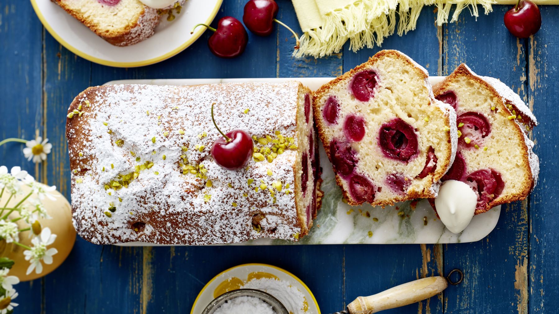 Saftiger Joghurt-Kirschkuchen in Scheiben geschnitten, mit ganzen Kirschen, gehackten Pistazien und Puderzucker bestreut, serviert auf einer Marmorplatte, daneben frische Kirschen und ein Klecks Sahne auf rustikalem, blauem Holztisch.