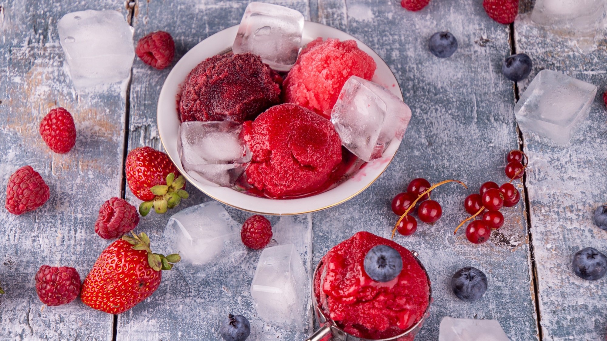 Drei Kugeln Beerensorbet mit Eiswürfeln in einer Schale, umgeben von frischen Erdbeeren, Himbeeren und Blaubeeren.