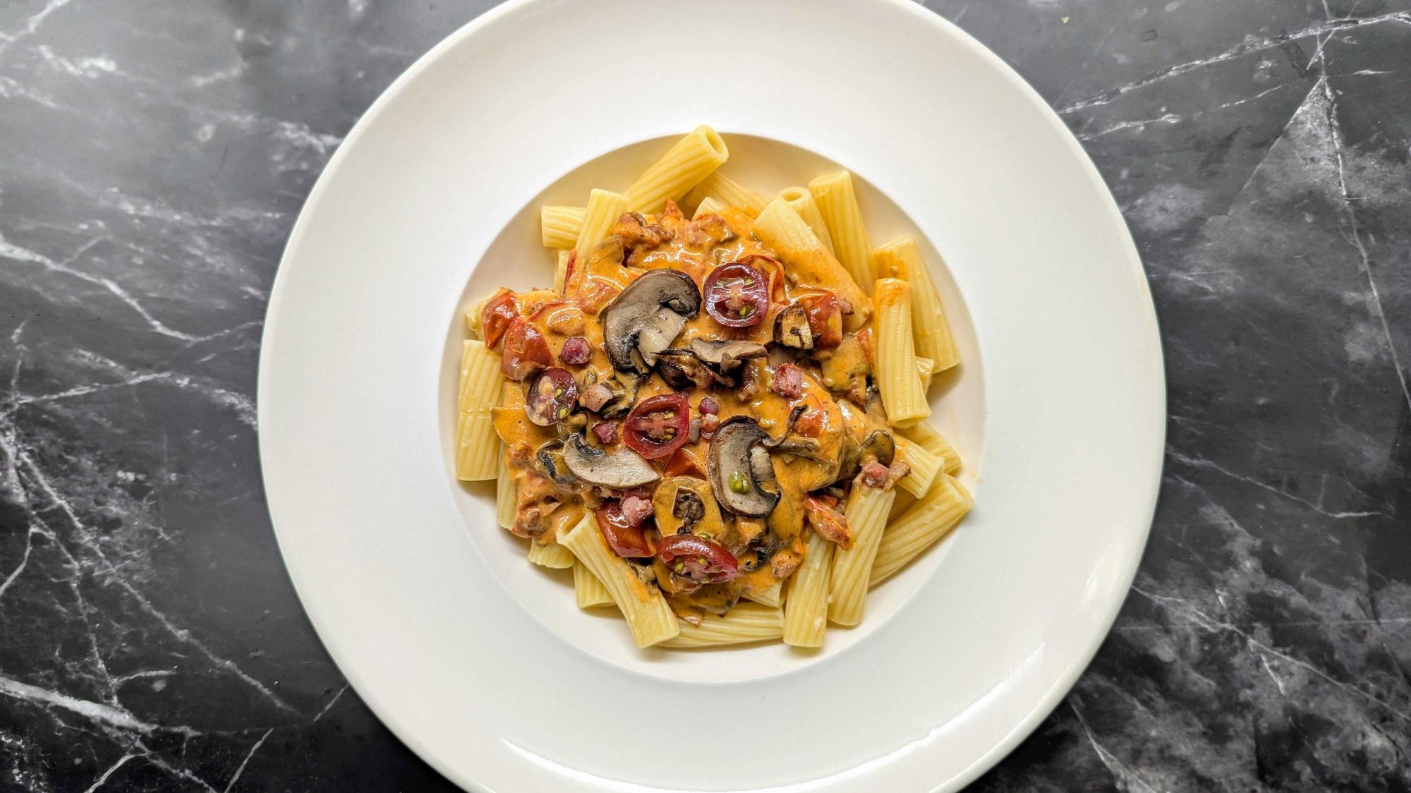 Cubana-Pasta auf weißem Teller: Pasta mit Mascarpone-Tomaten-Sauce, Champignons und Pancetta auf dunklem Marmortisch angerichtet.