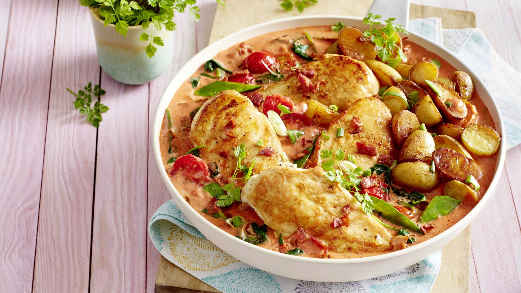 Toskanische Hähnchenpfanne mit goldbraun gebratenen Hähnchenbrustfilets, knusprigen Kartoffelhälften, Zuckerschoten, Spinat und Kirschtomaten in cremiger Tomatensauce, garniert mit frischen Kräutern, serviert in einer weißen Schale auf hellem Holztisch.