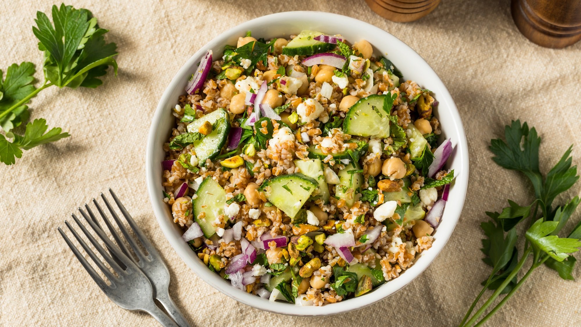 Bunter Bulgur-Salat mit Kichererbsen, Gurke, roten Zwiebeln, Minze, Feta und Pistazien in weißer Schale auf Leinentuch.