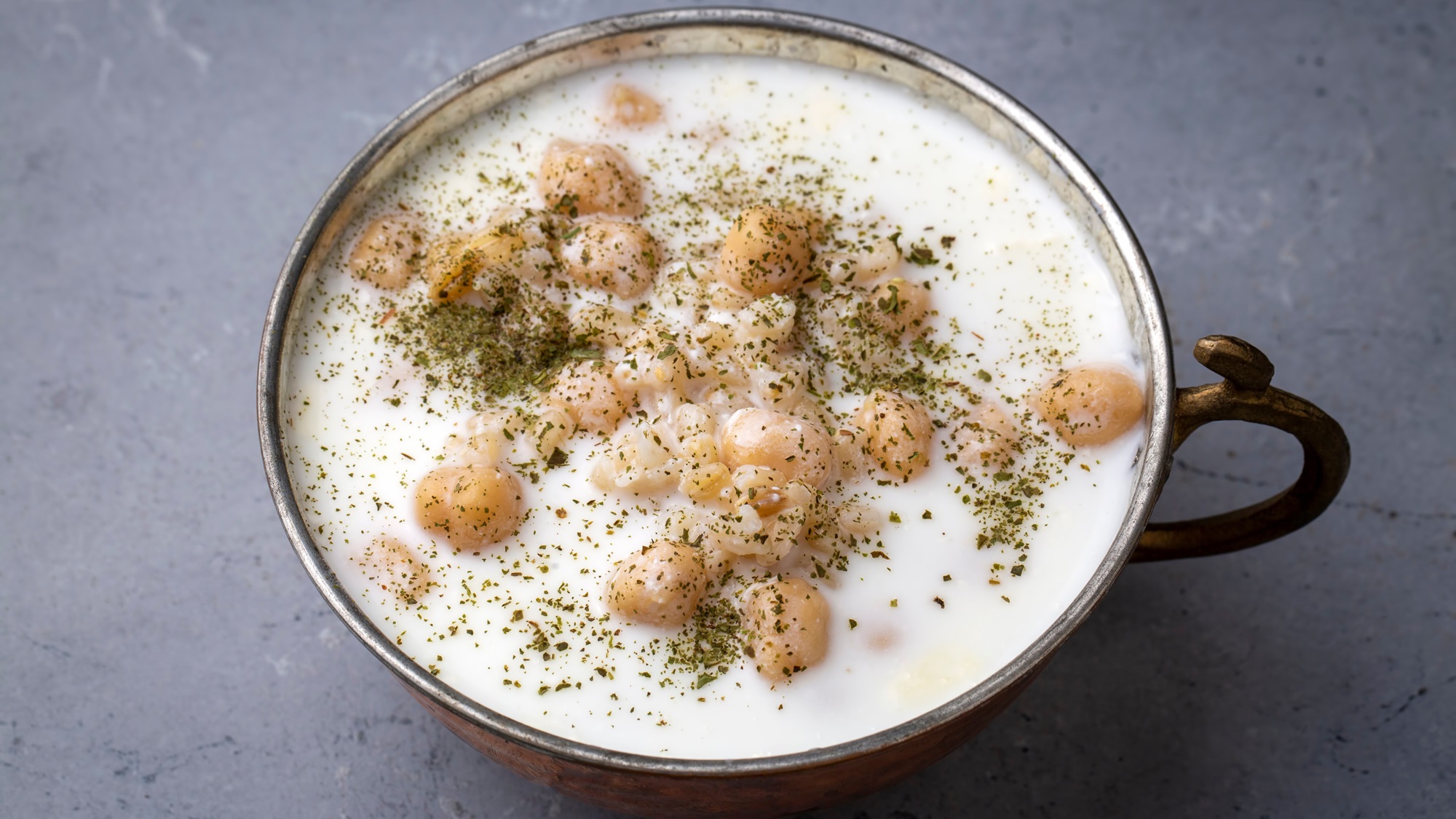 Cremige Joghurtsuppe mit Kichererbsen, Graupen und Minze in einer bronzefarbenen Schale auf grauem Steinuntergrund.