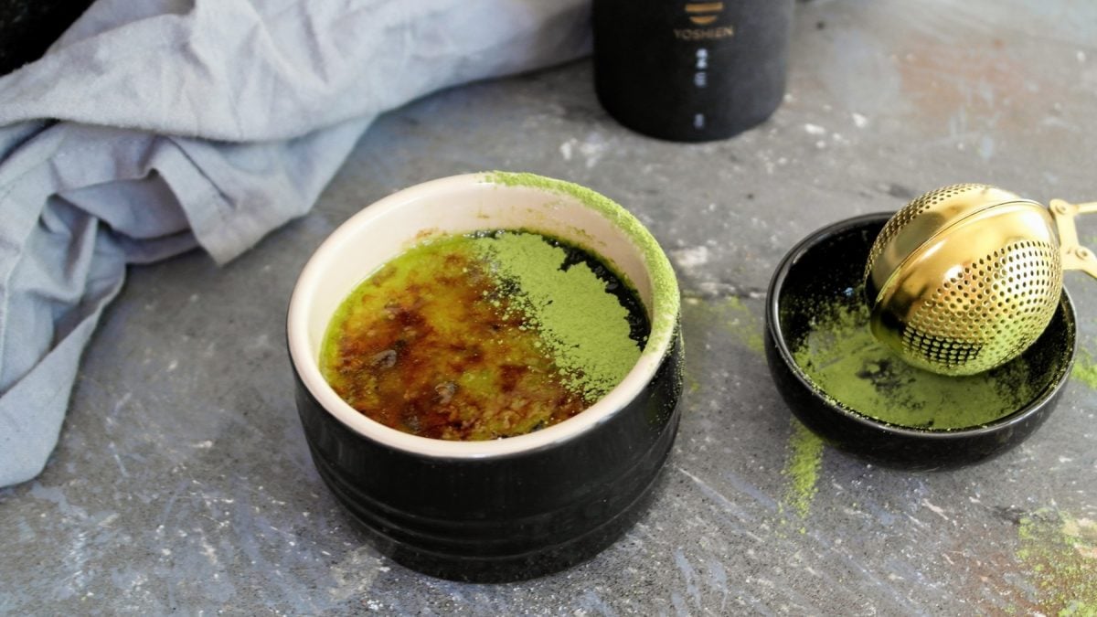 Ein schwarzes Schälchen mit der grünen Crème Brûlée auf grauem Tisch mit Matchapulver. Alles von schräg oben fotografiert.
