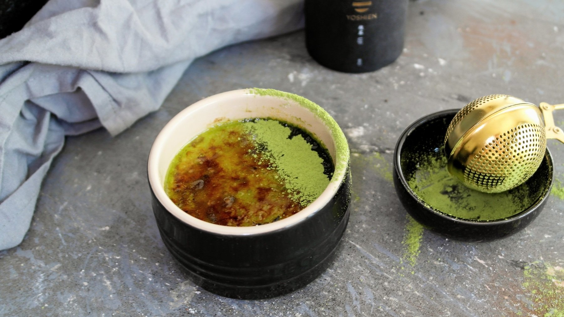 Ein schwarzes Schälchen mit der grünen Crème Brûlée auf grauem Tisch mit Matchapulver. Alles von schräg oben fotografiert.