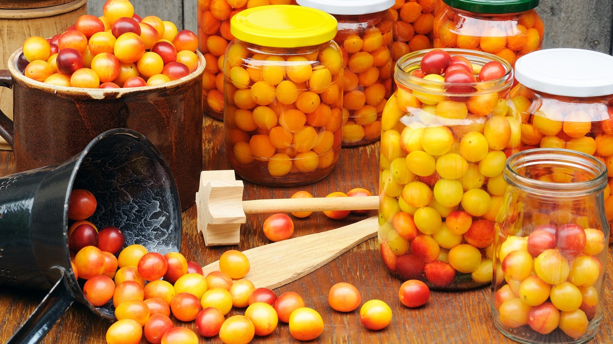 Ein übervoller schwarzer Eimer kippt bunte Mirabellen auf einen Tisch, umgeben von Einmachgläsern mit leuchtendem Obst.