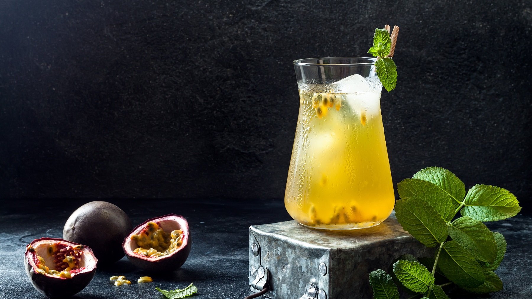 Ein großes Glas mit dem Passionsfrucht-Eistee mit dunklem Hintergrund. Daneben halbierte Passionsfrüchte und Minze.