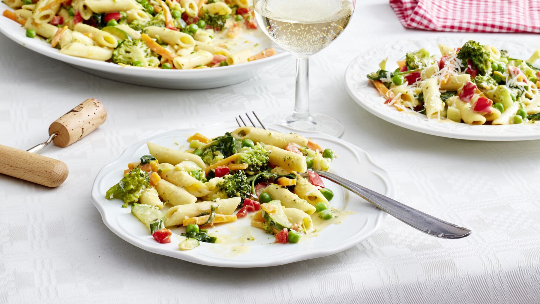 Ein weißer Teller mit Penne Primavera im One-Pot-Style, bunt gemischt mit Brokkoli, Erbsen, Zucchini, Paprika und Möhren in einer cremigen Sauce, daneben ein Glas Weißwein und weitere Teller mit Pasta auf einem hellen, gedeckten Tisch.